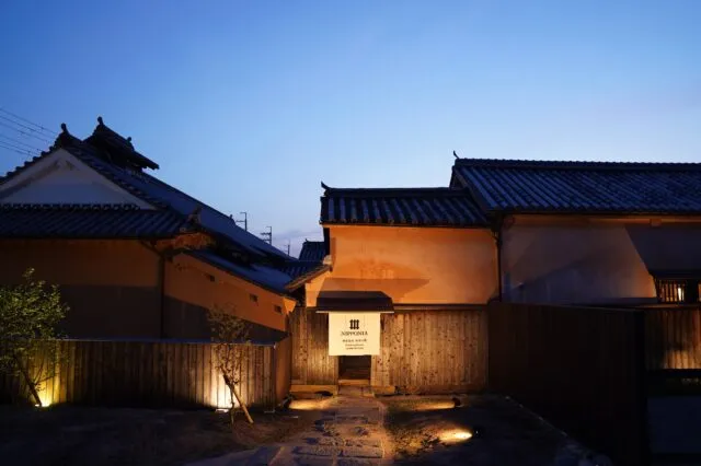 夕暮れの蔵書の館の外観。瓦屋根の蔵と門に明かりがともる