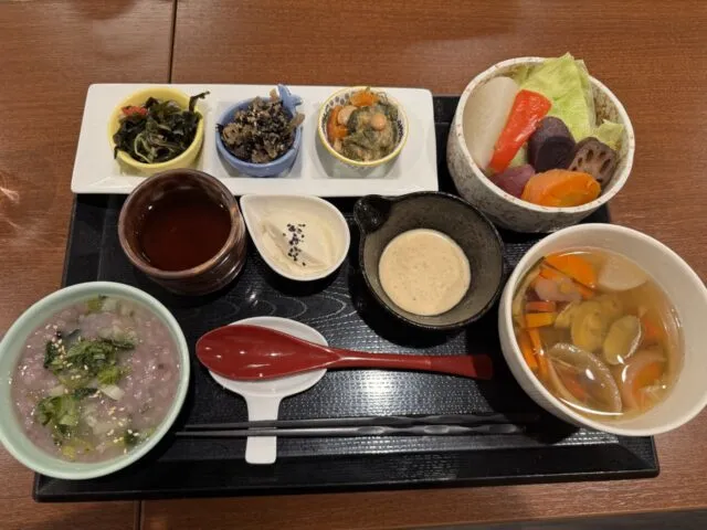 黒い盆に薬膳粥、野菜、3種の小鉢、豆腐、貝の入った汁物。木のテーブルと赤いれんげ。（2025年1月7日撮影）