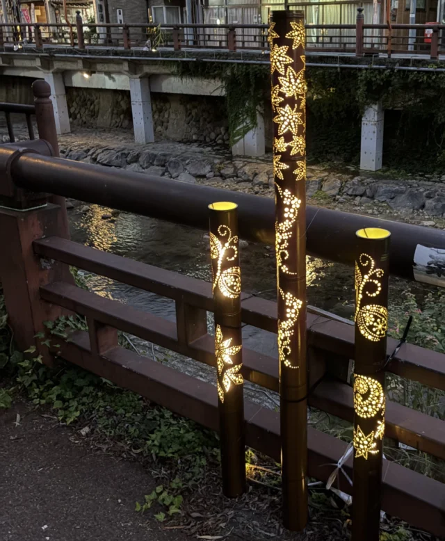 春来川沿いの柵に沿って立つ3本の竹灯籠。穴から漏れる光が花や曲線の模様を描く。