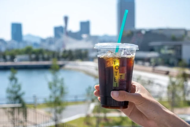 TOTTEIの公式店舗写真。港を背景にアイスコーヒーを持つ様子