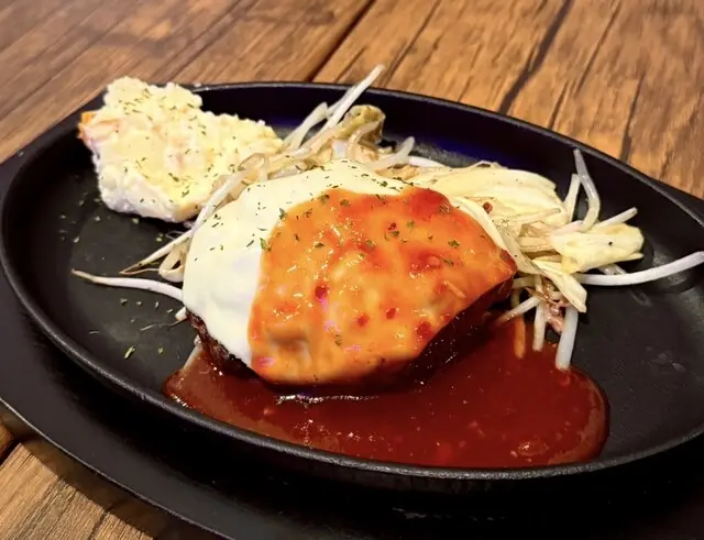 店舗の料理メニュー掲載写真。ハンバーグにチーズと赤いソースを重ね、鉄板に盛り付けた提供例