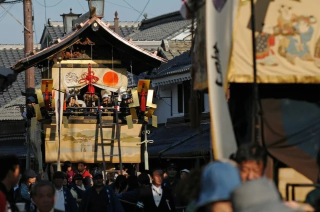 城下町の瓦屋根を背景に進む、金色の装飾と幕を備えた春日神社秋祭りの鉾山
