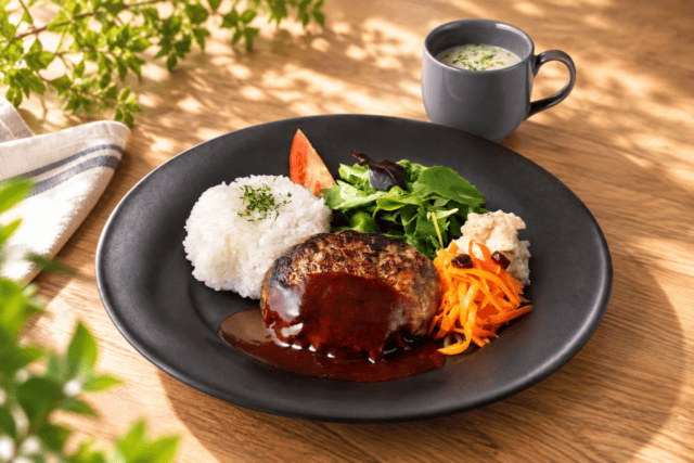 まねき食品の発表資料に掲載されたハンバーグプレート。黒い皿にハンバーグ、ライス、サラダが盛られている
