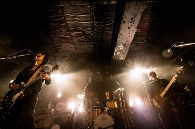 神戸VARIT.公演案内に掲載されたTHE GROOVERSの演奏紹介写真。ギター、ベース、ドラムとステージ照明