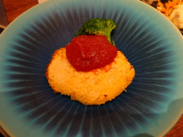 青い皿に平たい円形のカニクリームコロッケを載せ、赤いソースとブロッコリーを添えた料理