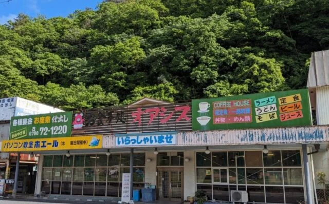 ドライブインオアシスの外観。横長の建物に赤いオアシスの看板、大きな窓、背後の緑の山