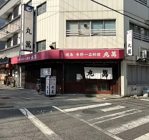 丸萬の掲載外観。白いのれん、赤い庇、淡い色の外壁がある角地の入口