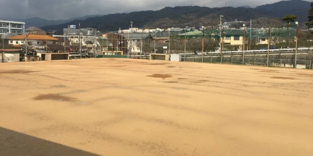 会場のシンコースポーツグラウンド。砂地の運動場を防球ネットが囲み、周囲に建物と山並みが見える。