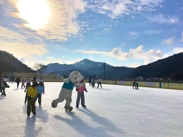 円山川公苑の屋外アイススケートリンク。周囲の柵、木々、背後の山が見える施設紹介写真。
