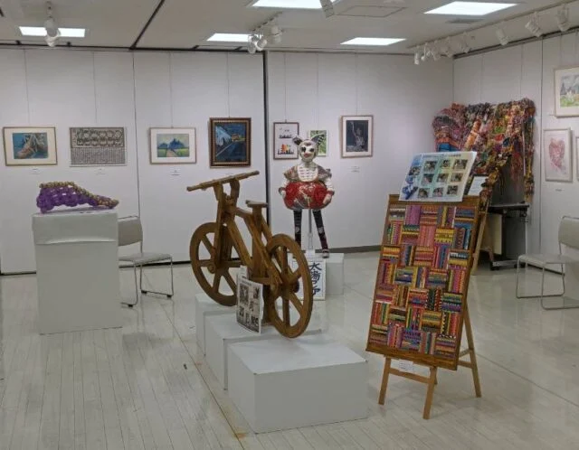 市公式掲載の展示資料写真。額装絵画と木の自転車型作品、色彩のある立体作品が並ぶ展示例。