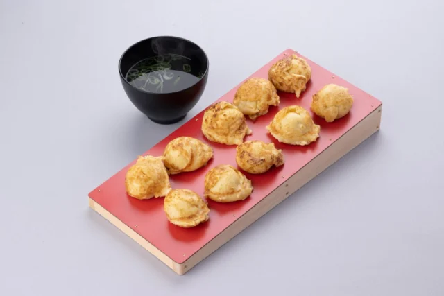 公式掲載の明石焼き風たこ焼き。焼き色のついた黄色い生地10個が赤い板に並び、別の器にだしが添えられている