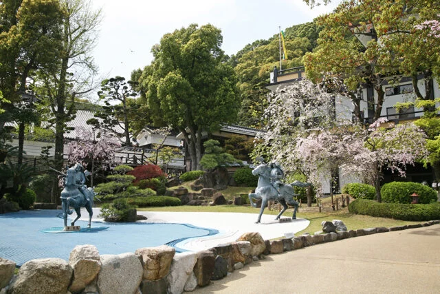 須磨寺公式の源平の庭の掲載写真。桜と緑を背景に2騎の像が向かい合い、手前の石の縁で庭と歩道が区切られています。撮影日は未確認