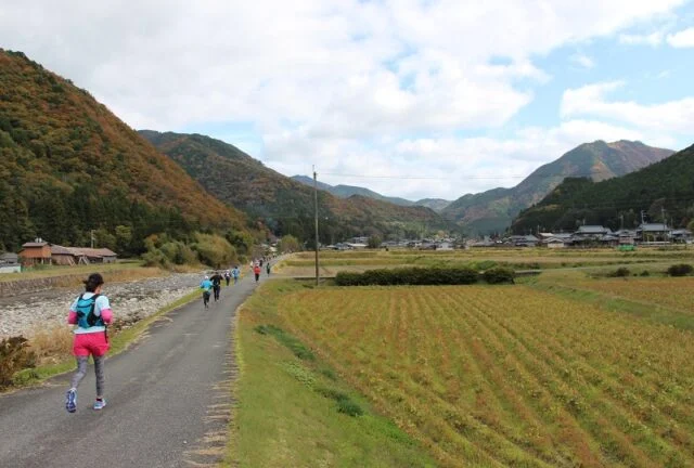 川沿いの舗装路と田畑、山々の間を進む過去の参加者。公式掲載写真、撮影年不明
