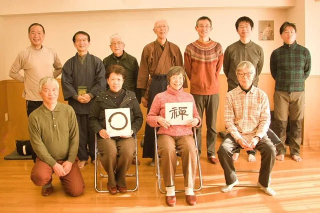 市が掲載する坐禅塾の参加者写真。木の床の部屋に椅子に座る人と立つ人が並ぶ。